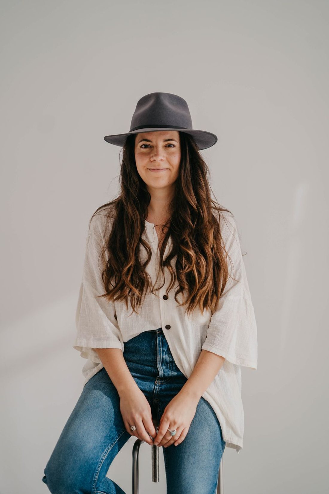 Angels Perez sitting on a stool in a cream shirt, jeans and a grey hat, looking at the camera