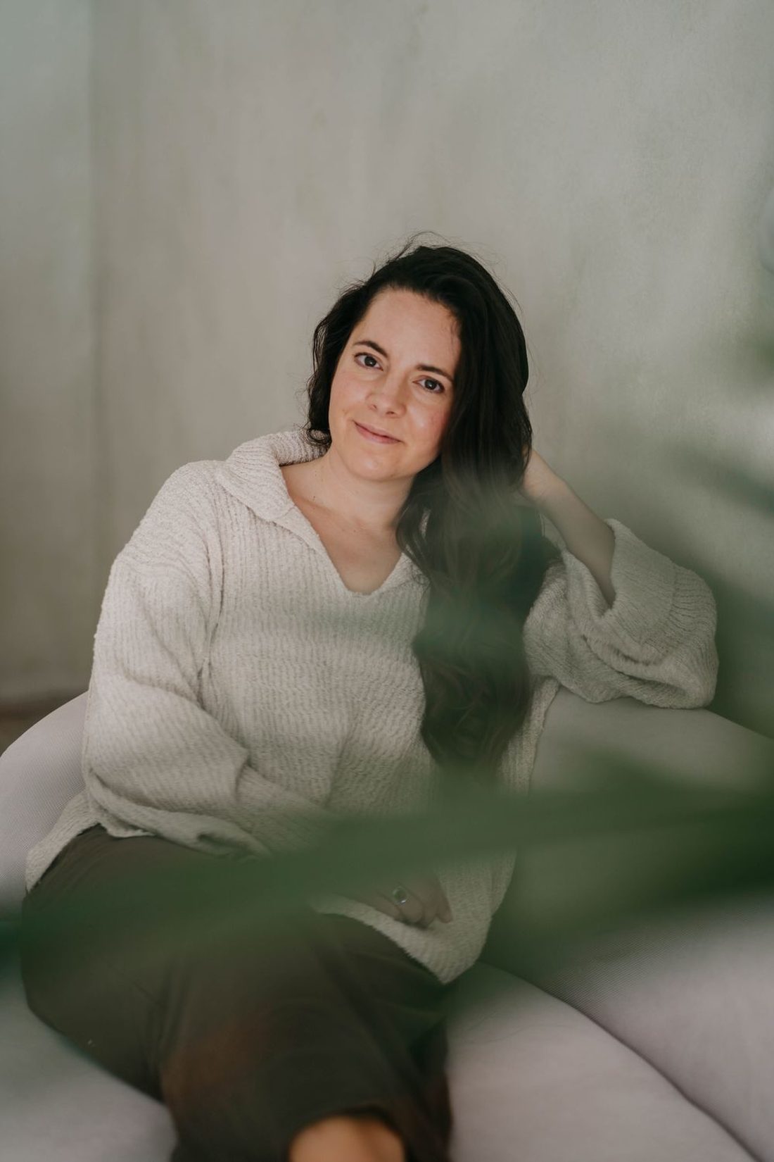 Angels Perez resting on a sofa in a cream jumper, seen through green leaves, with a soft, thoughtful look