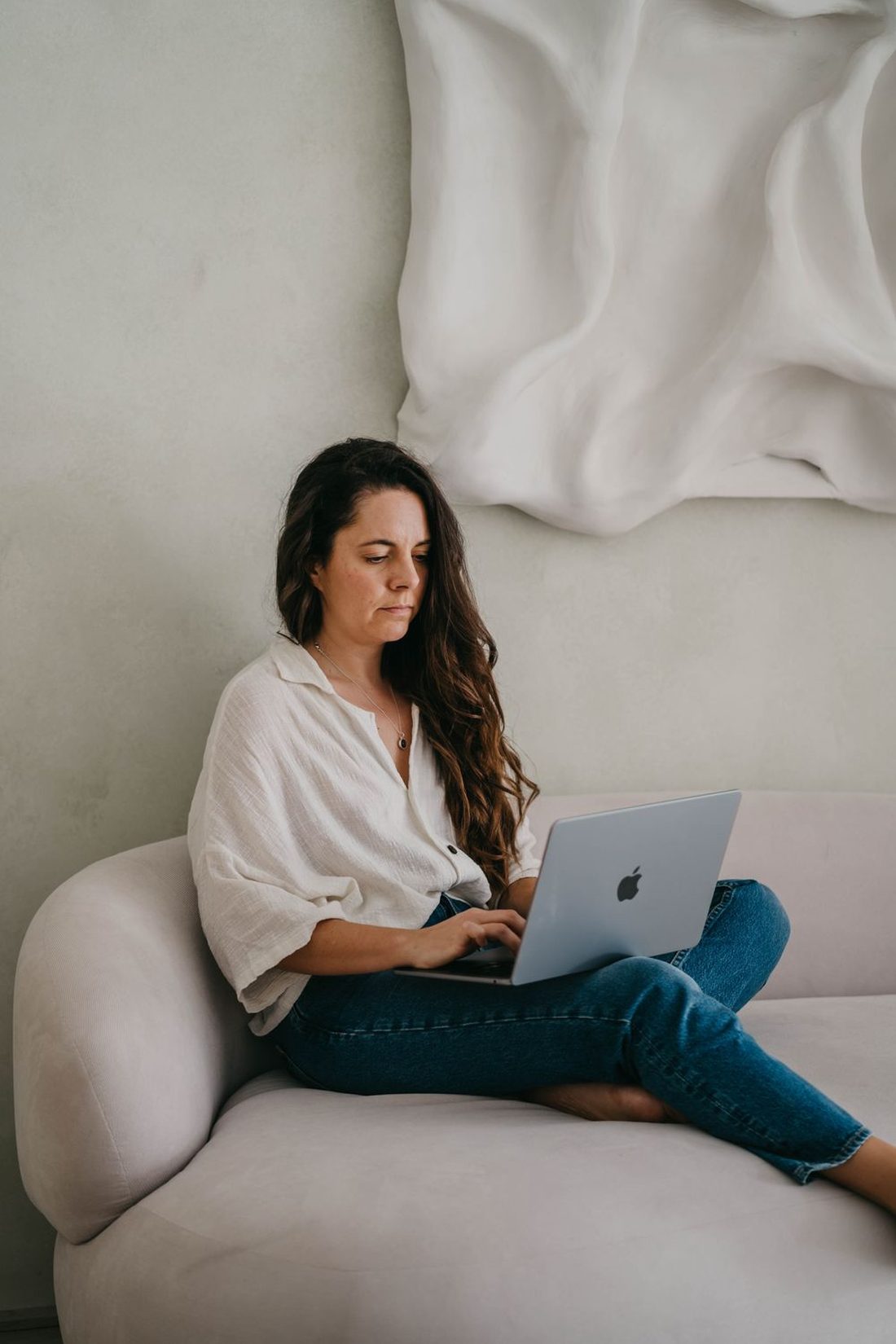 Angels Perez sitting on a cream sofa in jeans, reading on her laptop